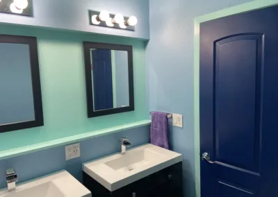 A modern bathroom with teal walls, navy blue door, a black vanity with a white sink, and two framed mirrors under bright lights.